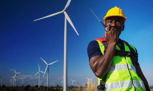 A person in a vest and hard hat in front of a wind turbine